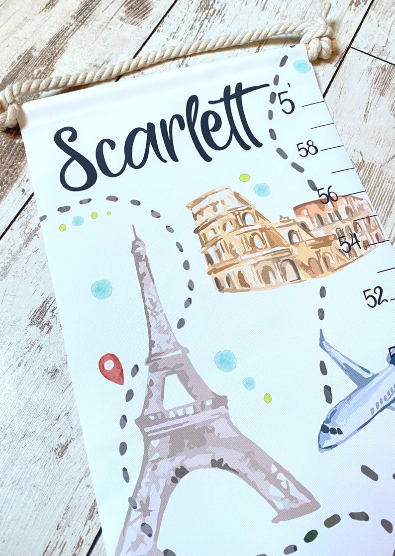 Puede incluir: Un medidor de altura de tela blanca para colgar con una cuerda, que presenta el nombre Scarlett en letra negra, ilustraciones de la Torre Eiffel y el Coliseo, un avi&oacute;n azul y una escala vertical marcada en cm del 52 al 60.