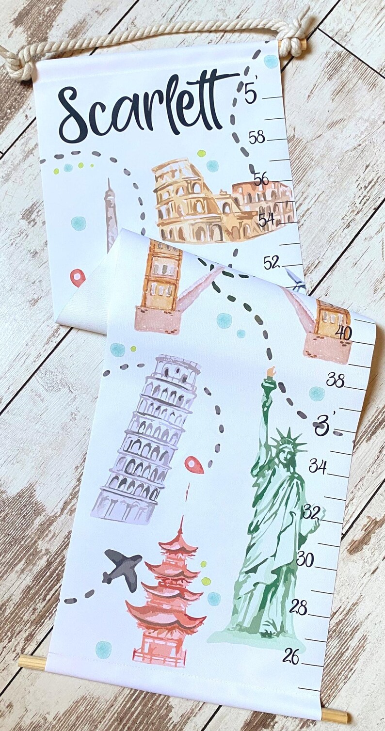 Puede incluir: Un medidor de altura de lona blanca que cuelga de una varilla de madera con una cuerda. Presenta el nombre Scarlett, una ruta de viaje punteada e ilustraciones en acuarela del Coliseo, la Torre de Pisa, la Estatua de la Libertad y una pagoda.