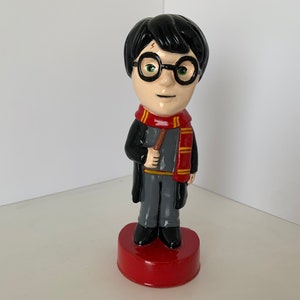 Puede incluir: Una figurita de cerámica de Harry Potter, con una túnica negra, una bufanda roja y dorada y gafas redondas. Lleva una varita mágica en la mano derecha y tiene una cicatriz en forma de rayo en la frente.
