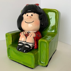 Puede incluir: Una figurita de cerámica de una niña con cabello negro sentada en un sillón verde. La niña lleva un vestido rojo y zapatos negros.