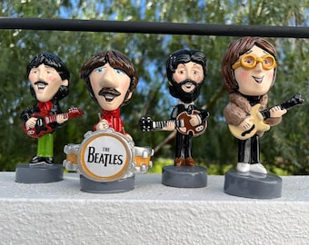 May include: Four small figurines of the Beatles, each with a unique hairstyle and facial features. The figures are wearing clothing and holding musical instruments. The figure in the center is playing a drum set with a drum that reads "The Beatles".