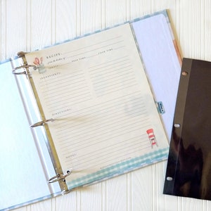 May include: A blue and white recipe binder with a clear plastic sheet protector. The binder has a silver ring mechanism and a white background with a blue and white checkered pattern. The sheet protector is blank and has three holes for the binder.