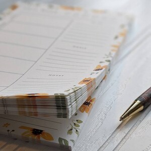 May include: A stack of blank notepads with a floral design featuring yellow sunflowers and green leaves. The notepads are stacked on a white surface with a gold pen lying next to them.