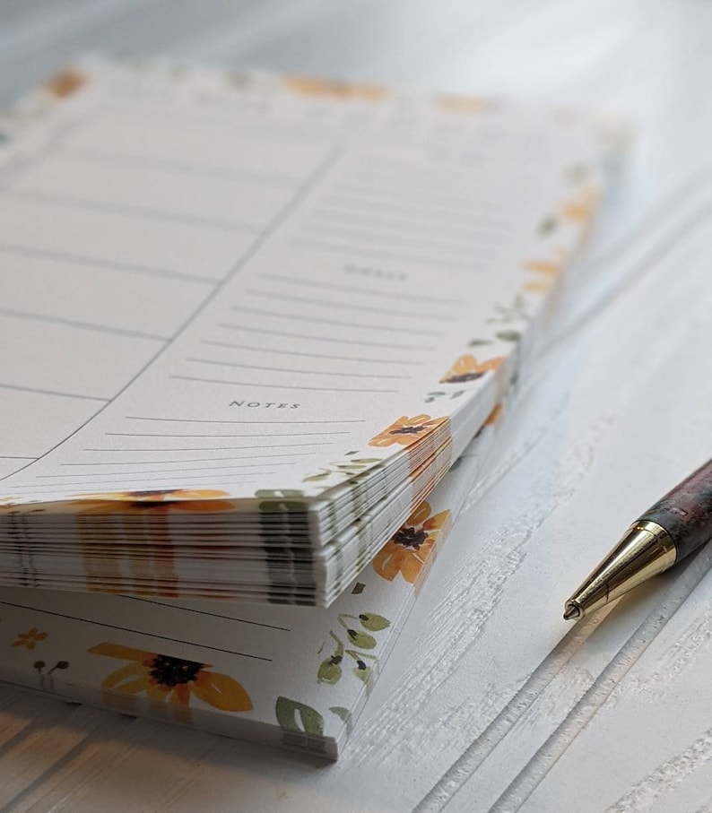 May include: A stack of blank notepads with a floral design featuring yellow sunflowers and green leaves. The notepads are stacked on a white surface with a gold pen lying next to them.