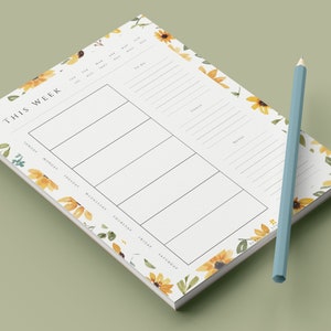 May include: A weekly planner notepad with a sunflower and floral design. The notepad has a white background with black text and lines. The text "This Week" is at the top of the notepad. The days of the week are listed below the text. The notepad is surrounded by a border of sunflowers and floral designs.