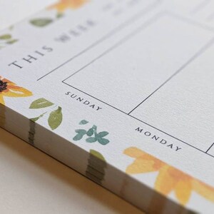 May include: A white notepad with a floral design featuring sunflowers and green leaves. The notepad has a weekly planner layout with the days of the week labeled: Sunday, Monday, Tuesday.