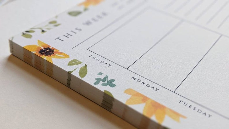 May include: A white notepad with a floral design featuring sunflowers and green leaves. The notepad has a weekly planner layout with the days of the week labeled: Sunday, Monday, Tuesday.