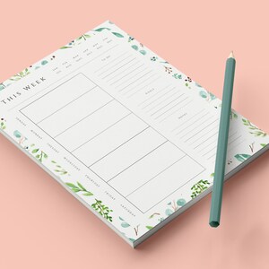 May include: A white weekly planner notepad with a floral design. The notepad has a section for each day of the week, a section for goals, and a section for notes. A green pencil is lying next to the notepad.