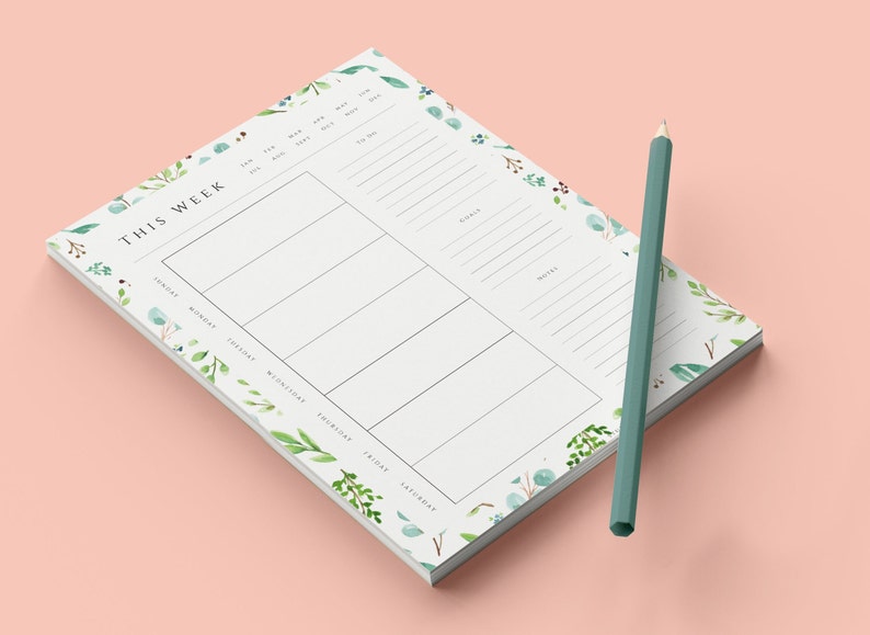 May include: A white weekly planner notepad with a floral design. The notepad has a section for each day of the week, a section for goals, and a section for notes. A green pencil is lying next to the notepad.