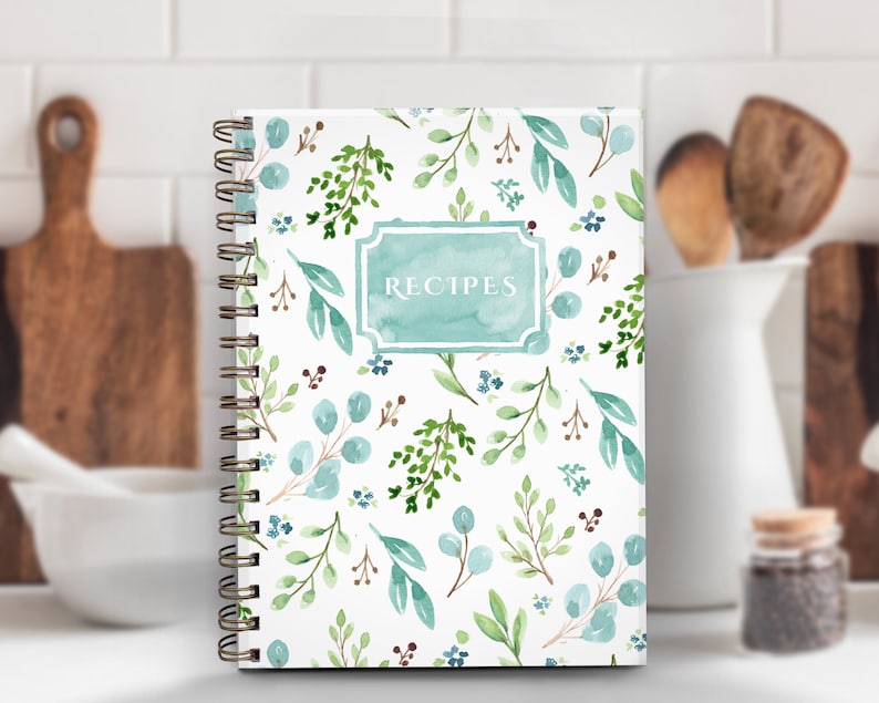 May include: A spiral-bound recipe journal with a white cover featuring a watercolor pattern of green and blue leaves and sprigs. The word "RECIPES" is in a teal frame. The journal is on a white surface.