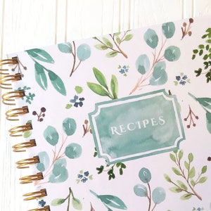 May include: A spiral-bound recipe book with a watercolor floral design. The cover features a white background with green and blue eucalyptus leaves and small blue flowers. The word "RECIPES" is in a teal frame.