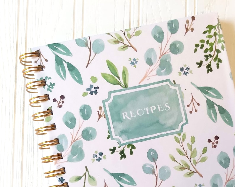 May include: A spiral-bound recipe book with a watercolor floral design. The cover features a white background with green and blue eucalyptus leaves and small blue flowers. The word "RECIPES" is in a teal frame.