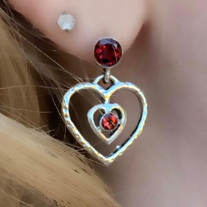 May include: Silver heart-shaped earrings with a red crystal in the center of each heart. The earrings have a delicate, layered design.