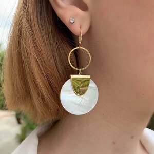May include: Gold hoop earrings with a white mother-of-pearl disc pendant. The disc has a gold accent at the top.