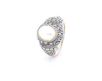 May include: A silver ring with a white pearl set in a detailed oval design. The ring is decorated with small, dark gray stones.