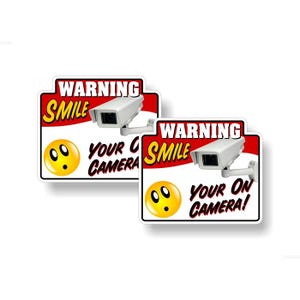 Puede incluir: Dos señales de advertencia rojas y blancas con el texto "WARNING SMILE YOUR ON CAMERA!" y una cara sonriente de dibujos animados. Cada señal presenta una cámara de seguridad blanca.