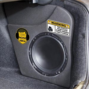 May include: A black car boot with a large speaker and a warning sticker that reads "WARNING THIS VEHICLE'S SOUND SYSTEM IS MORE POWERFUL THAN IT'S ENGINE". There is also a sticker that reads "LOUD NOISE AREA" with a headphone icon.