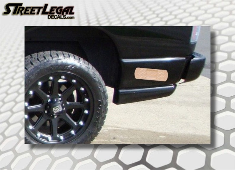 May include: A black pickup truck with a black wheel and a brown band-aid sticker on the rear bumper. The truck is parked on a gray concrete surface.
