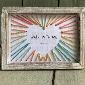 Può includere: Un'opera d'arte incorniciata con un design a forma di cuore realizzato con fili colorati. Il testo "WALK WITH ME" e "MOSES 6:34" è centrato all'interno del cuore. La cornice è in legno chiaro invecchiato.