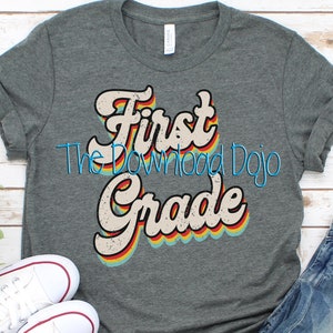 May include: A gray t-shirt with a retro-style graphic that says "First Grade" in rainbow colors. The graphic has a distressed look and is centered on the shirt.