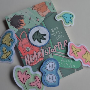 Op de afbeelding: Een exemplaar van "Heartstopper Volume 1" van Alice Oseman met verschillende bladvormige viltpatches. De boekomslag toont twee figuren en de tekst "Boy Meets Boy". De patches zijn in verschillende kleuren.