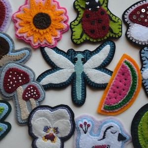 Op de afbeelding: Een verzameling kleurrijke viltapplicaties in verschillende vormen, waaronder een zonnebloem, lieveheersbeestje, libelle, paddenstoel, watermeloen, bij, slak en blad. Elke applicatie is met de hand gestikt met gedetailleerde accenten.