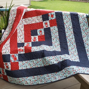 Può includere: Una trapunta rossa, bianca e blu con un motivo floreale. La trapunta è realizzata con quadrati e rettangoli di tessuto e ha un bordo blu scuro.