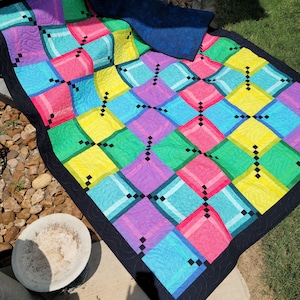 May include: A colorful quilt with a geometric pattern of squares and diamonds in shades of pink, blue, green, and yellow. The quilt is edged with black fabric and has a black backing.