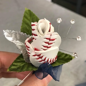 May include: A white and red baseball-themed rose buttonhole with green leaves and a blue ribbon. The rose is made from a baseball, with the laces visible.