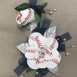 May include: Two baseball-themed flower arrangements with white baseballs, red stitching, and navy blue ribbon. The larger arrangement has a silver leaf and rhinestones.