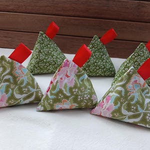 May include: Six handmade fabric triangle sachets. The sachets are green with floral and dot patterns, and each has a red ribbon. The sachets are arranged on a white surface with a wooden background.