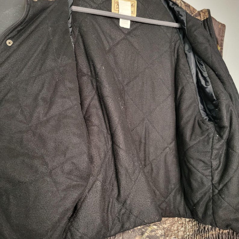 May include: Open view of a camouflage hunting jacket interior. The jacket is black with a quilted pattern. The interior lining is visible, along with the jacket's collar and a portion of the exterior camouflage fabric.