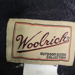 May include: Close-up of a rectangular beige label with the word "Woolrich" in a cursive font. The label has a red and black border and the text "OUTDOOR GUIDE COLLECTION". The label is attached to a dark gray fabric.