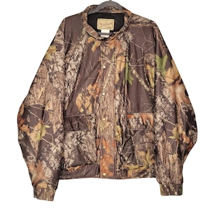 May include: A camouflage jacket with a button-up front, two front pockets, and a black collar. The jacket features a pattern of brown, green, and tan leaves and branches. The brand name "Woolrich" is visible on the collar.