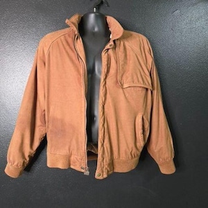 Könnte beinhalten: Braune Wildlederjacke mit Reißverschluss, zwei Vordertaschen und einem Kragen. Die Jacke wird auf einer schwarzen Schaufensterpuppe präsentiert und hat gerippte Bündchen und Saum. Die Jacke scheint im Vintage-Stil zu sein.