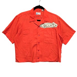 May include: A bright orange short-sleeved button-down shirt with a collar and two front chest pockets. A white banner patch is stitched onto the upper right chest area, featuring the black hand-lettered text Piece of Shit in a stylized font.