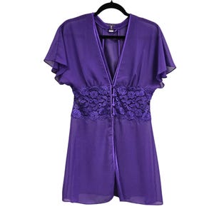 May include: A sheer purple robe with short, flutter sleeves and a V-neckline. The robe features a decorative lace panel at the waist and a button closure. The garment is hanging on a black hanger.