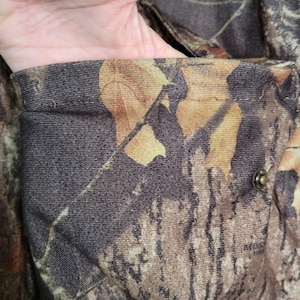May include: Close-up of a camouflage jacket pocket. The fabric features a pattern of brown, green, and tan leaf and branch designs. A metal snap is visible on the pocket's edge, and the pocket is open.