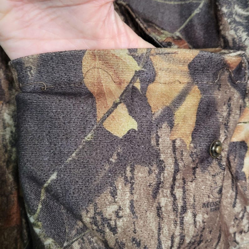 May include: Close-up of a camouflage jacket pocket. The fabric features a pattern of brown, green, and tan leaf and branch designs. A metal snap is visible on the pocket's edge, and the pocket is open.
