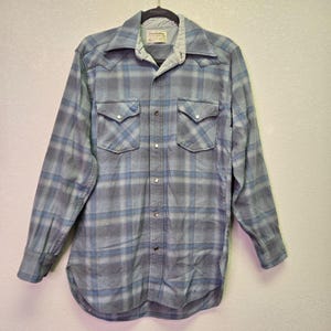 May include: A long-sleeved, button-down shirt in a blue and gray plaid pattern. The shirt has two chest pockets with pointed flaps and silver-colored buttons. The collar and cuffs are a solid light blue color.