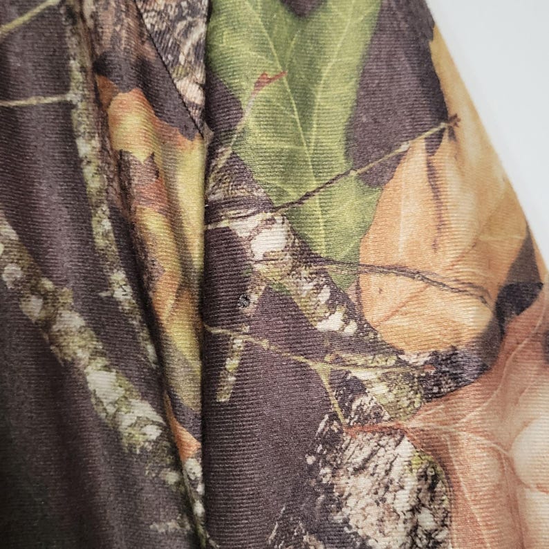 May include: Close-up of a camouflage pattern fabric. The design features a mix of brown, green, and tan colors, resembling leaves and branches. The fabric appears to be a durable material, suitable for outdoor wear.