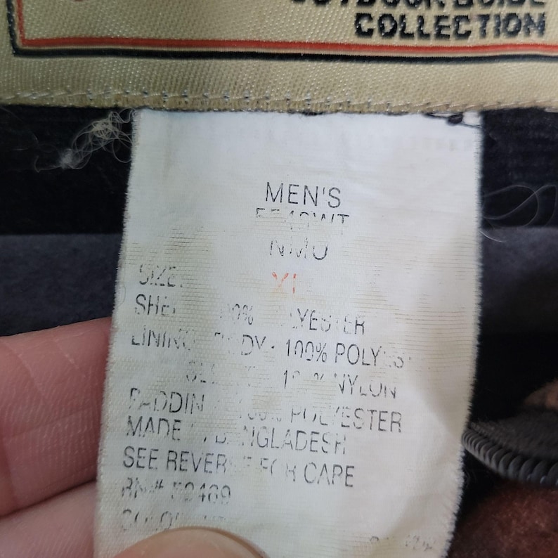 May include: Close-up of a white clothing tag with black text. The tag reads "MEN'S" and includes details about the garment's size, shell, lining, and padding composition. The tag also indicates the item was made in Bangladesh.