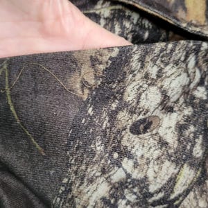 May include: Close-up of camouflage fabric with a detailed pattern of brown, green, and tan hues. The fabric has a textured appearance, with visible stitching along the edge. The overall design mimics a forest environment.