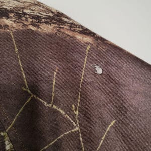 May include: Close-up of a section of camouflage fabric. The fabric is primarily brown with a pattern of tan and green. There is a small, light-colored spot on the fabric. The stitching is visible in a lighter color.