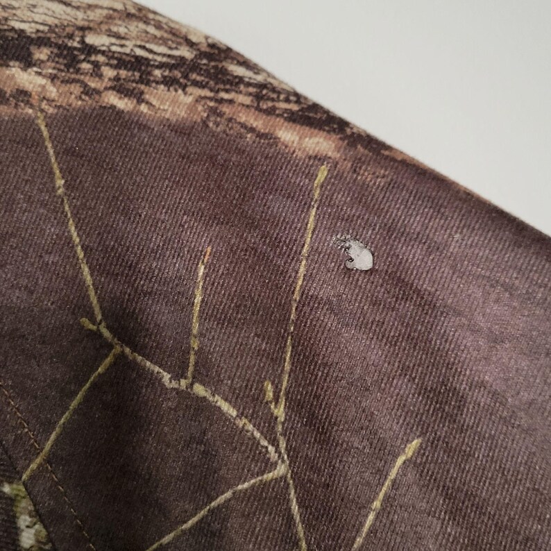 May include: Close-up of a section of camouflage fabric. The fabric is primarily brown with a pattern of tan and green. There is a small, light-colored spot on the fabric. The stitching is visible in a lighter color.