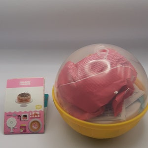 May include: A yellow plastic sphere container with a clear dome lid holding pink wrapped items, accompanied by a small rectangular instruction card labeled Tiramisu and a circular white scalloped paper disc with a central M logo.