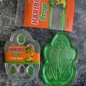 Può includere: Una vista dall'alto presenta uno stampo per caramelle in plastica trasparente e uno stampo a forma di rana in plastica verde, entrambi a tema rana. È visibile anche una confezione di caramelle Haribo Frogs. La confezione di caramelle è arancione con il logo Haribo e la grafica delle rane.