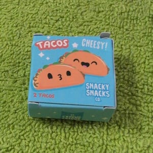 Op de afbeelding: Een kleine, lichtblauwe doos met de woorden "TACOS" en "Cheesy!" erop gedrukt. De doos bevat illustraties van twee lachende taco's. De tekst "2 TACOS" en "SNACKY SNACKS CO." zijn ook zichtbaar.