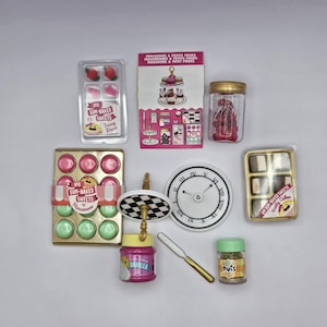 Pode incluir: Uma coleção de suprimentos de confeitaria em miniatura, incluindo um relógio, um suporte, um frasco de doces vermelhos, um frasco de extrato de baunilha, um frasco de nozes, uma faca e duas embalagens de macarons. As embalagens são rotuladas como "Les Sun-Baked Sweets" e apresentam uma combinação de cores rosa e verde.