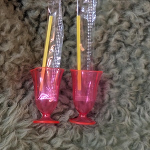 May include: Two pink plastic goblets with red bases, each containing a yellow straw and a silver-coloured, flexible cleaning brush in a clear plastic wrapper. The goblets are on a textured, olive-green surface.